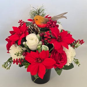 May include: A floral arrangement in a black container featuring bright red poinsettias, white roses, and white carnations. The display includes clusters of red berries, a brown pinecone, green foliage, and a small orange bird with grey wings.