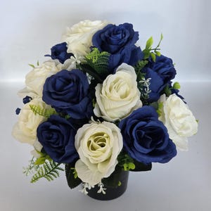 May include: A floral arrangement of navy blue and cream roses. The flowers are arranged in a black vase, with green foliage. A classic decorative bouquet.