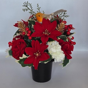 May include: A Christmas floral display in a black pot. It includes red poinsettias, roses, and berries, with white carnations and gold accents. A small bird ornament sits on top, adding a decorative touch.