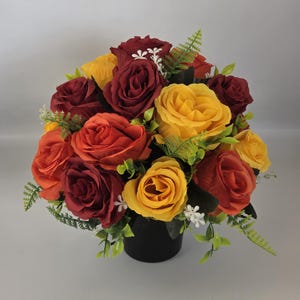 May include: A colourful floral arrangement featuring roses in shades of red, orange, and yellow. The roses are arranged in a black vase, accented with green foliage and small white flowers. A vibrant centrepiece.
