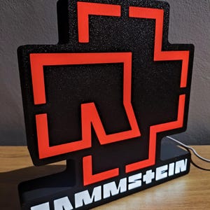 Puede incluir: Un letrero iluminado negro y rojo con el nombre de la banda "RAMMSTEIN". El dise&ntilde;o es una cruz roja sobre un fondo negro. El nombre de la banda est&aacute; escrito en blanco en la base.