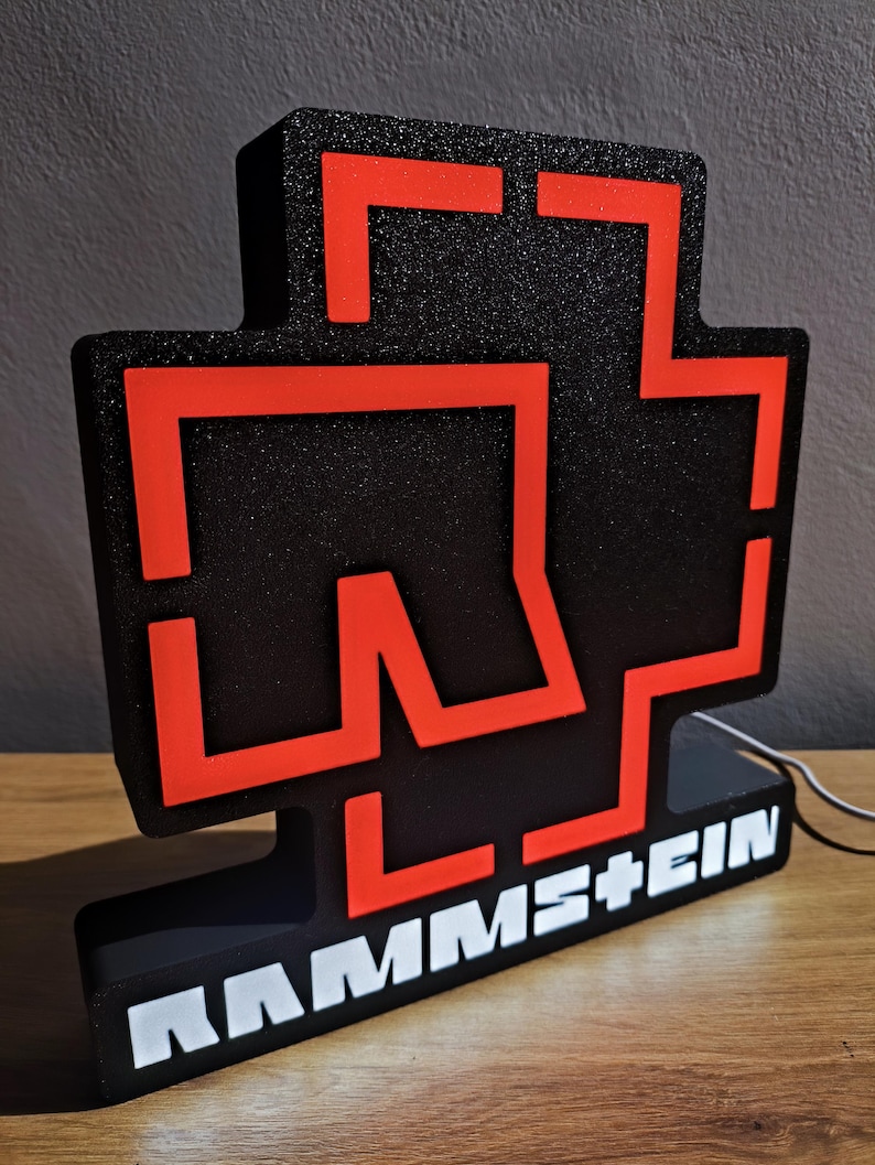 Puede incluir: Un letrero iluminado negro y rojo con el nombre de la banda "RAMMSTEIN". El dise&ntilde;o es una cruz roja sobre un fondo negro. El nombre de la banda est&aacute; escrito en blanco en la base.