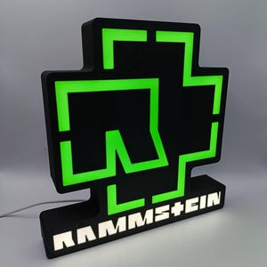 Puede incluir: Un letrero iluminado en negro y verde con el logotipo de la banda. El letrero tiene un dise&ntilde;o en forma de cruz con contornos de ne&oacute;n verde y el nombre de la banda, RAMMSTEIN, en letras blancas en la base. El letrero est&aacute; enchufado.