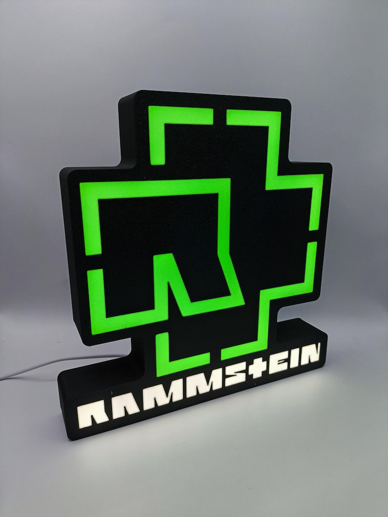 Puede incluir: Un letrero iluminado en negro y verde con el logotipo de la banda. El letrero tiene un dise&ntilde;o en forma de cruz con contornos de ne&oacute;n verde y el nombre de la banda, RAMMSTEIN, en letras blancas en la base. El letrero est&aacute; enchufado.