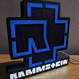 Puede incluir: Letrero iluminado en negro y azul con el nombre de la banda RAMMSTEIN. El letrero presenta un dise&ntilde;o en forma de cruz con detalles azules y el nombre de la banda en blanco. Colocado sobre una superficie de madera.