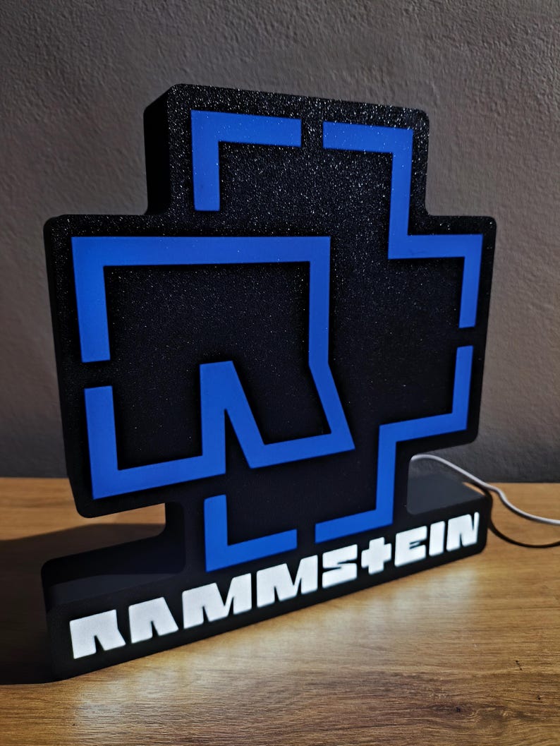 Puede incluir: Letrero iluminado en negro y azul con el nombre de la banda RAMMSTEIN. El letrero presenta un dise&ntilde;o en forma de cruz con detalles azules y el nombre de la banda en blanco. Colocado sobre una superficie de madera.