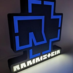 Puede incluir: Un letrero iluminado en negro y azul con el nombre de la banda "RAMMSTEIN" en letras blancas. El letrero tiene un dise&ntilde;o en forma de cruz con detalles azules y una base rectangular. La iluminaci&oacute;n interna crea un efecto brillante.