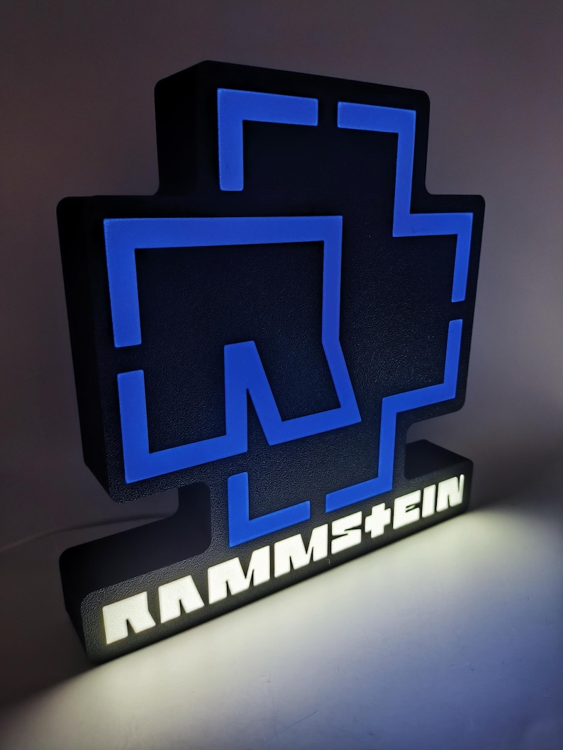 Puede incluir: Un letrero iluminado en negro y azul con el nombre de la banda "RAMMSTEIN" en letras blancas. El letrero tiene un dise&ntilde;o en forma de cruz con detalles azules y una base rectangular. La iluminaci&oacute;n interna crea un efecto brillante.