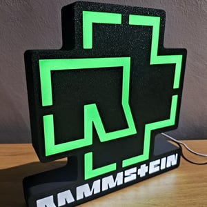 Puede incluir: Letrero luminoso negro y verde con el nombre de la banda RAMMSTEIN en letras blancas. El letrero tiene un dise&ntilde;o en forma de cruz con contornos verdes de estilo ne&oacute;n. Est&aacute; sobre una superficie de madera.