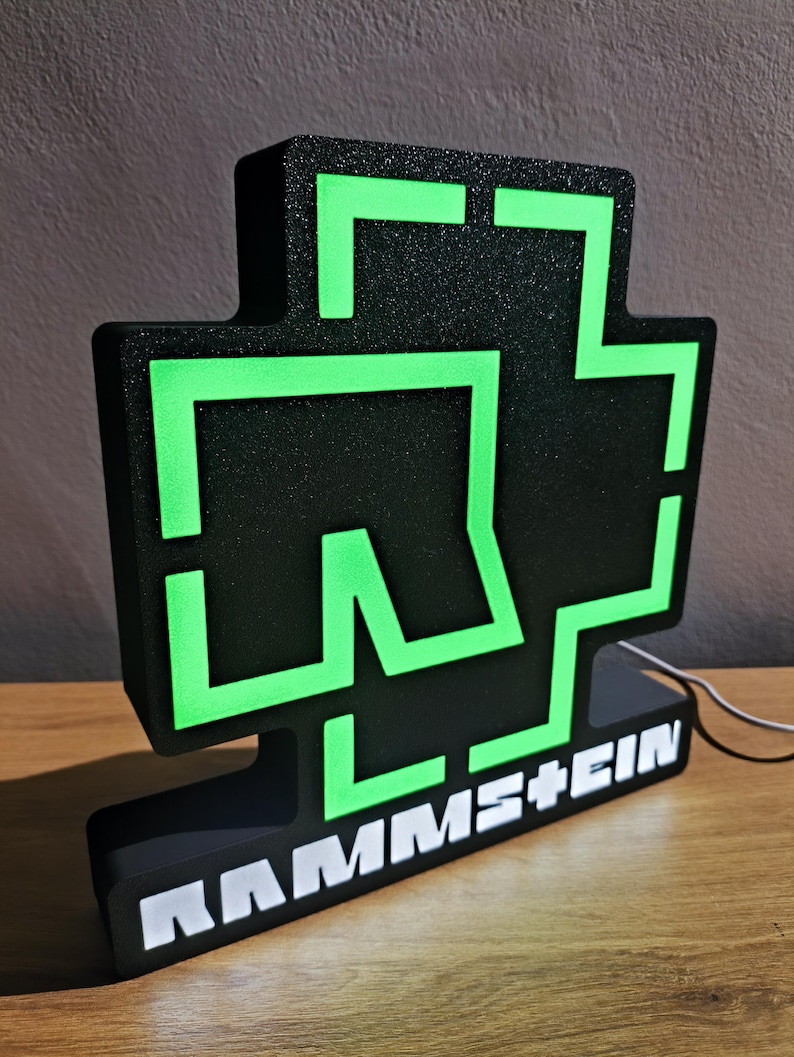 Puede incluir: Letrero luminoso negro y verde con el nombre de la banda RAMMSTEIN en letras blancas. El letrero tiene un dise&ntilde;o en forma de cruz con contornos verdes de estilo ne&oacute;n. Est&aacute; sobre una superficie de madera.