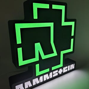 Puede incluir: Un letrero negro y verde iluminado con el logotipo de la banda. El logotipo es una cruz estilizada con el nombre de la banda, RAMMSTEIN, en letras blancas en la base. Los elementos verdes brillan sobre el fondo negro.