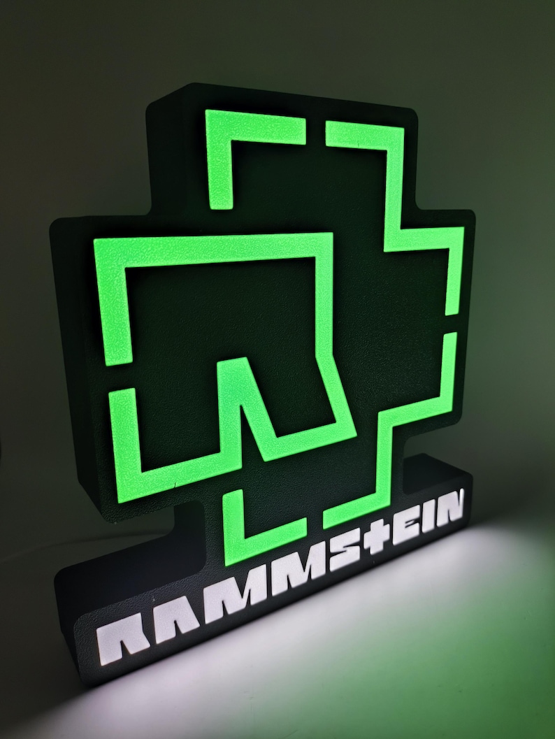 Puede incluir: Un letrero negro y verde iluminado con el logotipo de la banda. El logotipo es una cruz estilizada con el nombre de la banda, RAMMSTEIN, en letras blancas en la base. Los elementos verdes brillan sobre el fondo negro.