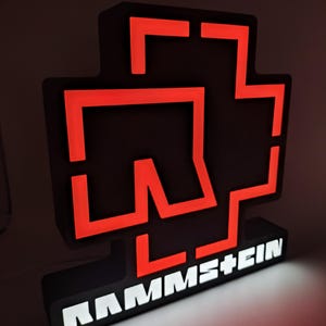 Puede incluir: Un letrero iluminado en negro y rojo con el nombre de la banda "RAMMSTEIN". El letrero presenta un logotipo distintivo con un contorno rojo y un fondo negro. El nombre de la banda se muestra en letras blancas iluminadas.