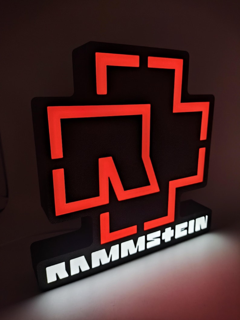 Puede incluir: Un letrero iluminado en negro y rojo con el nombre de la banda "RAMMSTEIN". El letrero presenta un logotipo distintivo con un contorno rojo y un fondo negro. El nombre de la banda se muestra en letras blancas iluminadas.