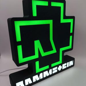 Puede incluir: Letrero luminoso negro y verde con el logotipo de Rammstein. El letrero tiene una superficie texturizada y el nombre de la banda se muestra en letras blancas en la base. Los elementos verdes brillan.