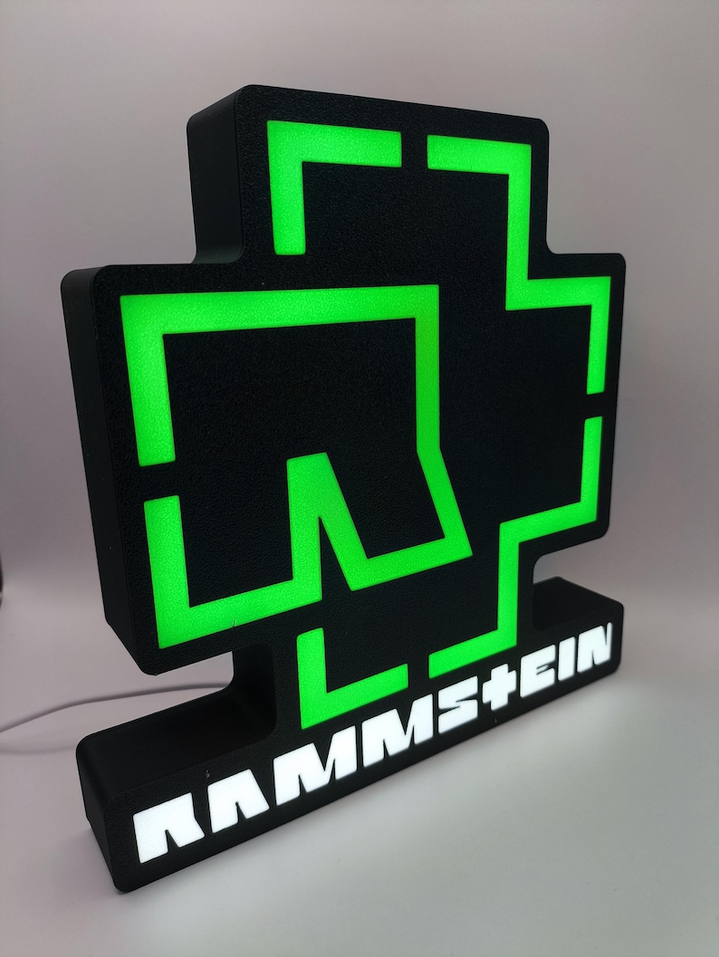 Puede incluir: Letrero luminoso negro y verde con el logotipo de Rammstein. El letrero tiene una superficie texturizada y el nombre de la banda se muestra en letras blancas en la base. Los elementos verdes brillan.