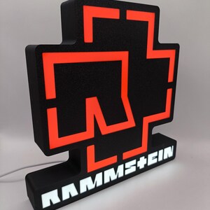 Puede incluir: Letrero luminoso negro y naranja con el logotipo de la banda. El letrero tiene una base negra con el nombre de la banda en letras blancas. El logotipo es una cruz estilizada con detalles en naranja.