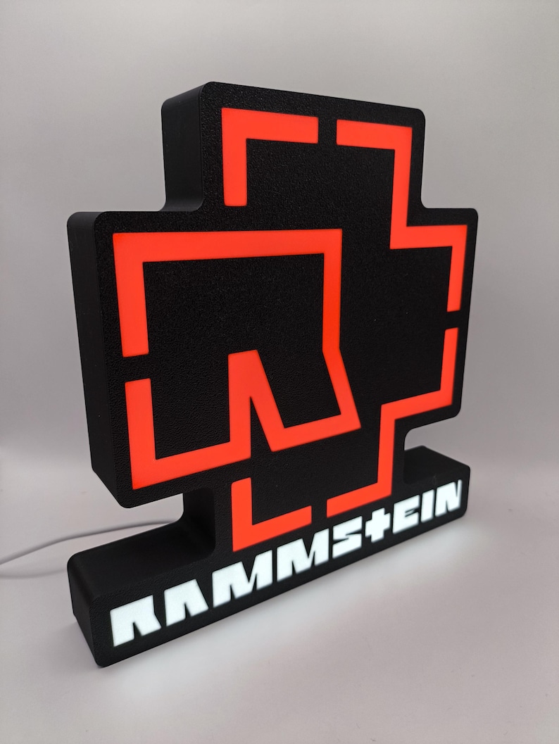 Puede incluir: Letrero luminoso negro y naranja con el logotipo de la banda. El letrero tiene una base negra con el nombre de la banda en letras blancas. El logotipo es una cruz estilizada con detalles en naranja.