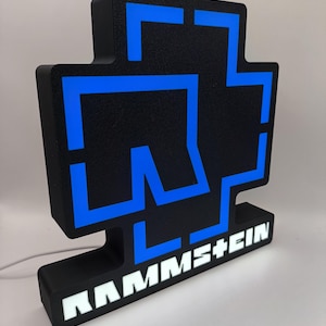 Puede incluir: Letrero luminoso negro y azul con el logotipo de la banda. El letrero tiene una superficie texturizada y el nombre de la banda, RAMM+EIN, se muestra en letras blancas en la base. Un cable de alimentaci&oacute;n blanco se extiende desde la parte posterior.