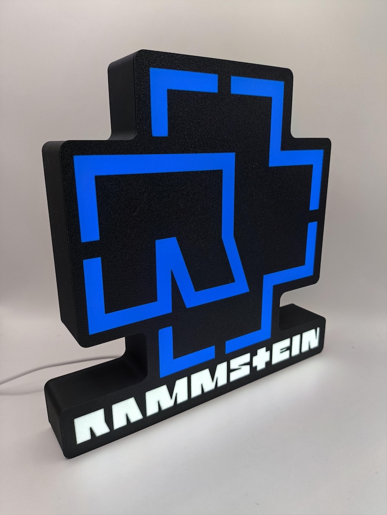 Puede incluir: Letrero luminoso negro y azul con el logotipo de la banda. El letrero tiene una superficie texturizada y el nombre de la banda, RAMM+EIN, se muestra en letras blancas en la base. Un cable de alimentaci&oacute;n blanco se extiende desde la parte posterior.
