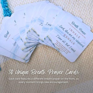 May include: A set of thirty square cards featuring a light blue watercolor ocean scene. Each card displays the text Breath Prayer, Inhale, Exhale, and specific scripture verses. The cards are fanned out, showing various prayer phrases printed in dark blue and brown ink.