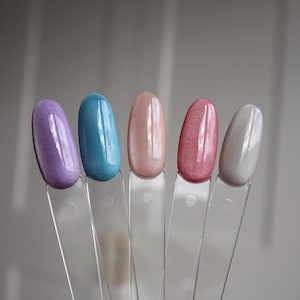 Könnte beinhalten: Fünf Nagellack-Farbmuster in einer Reihe, jeweils auf einem durchsichtigen Plastikstab. Die Farben sind Lila, Türkis, Hellrosa, Dunkelrosa und Silber. Der Nagellack hat ein glänzendes, metallisches Finish.