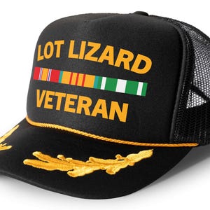 May include: Black trucker hat with a gold embroidered wreath and the text "LOT LIZARD VETERAN" in yellow. A row of colorful ribbons is above the text.
