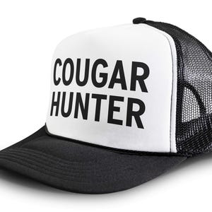 May include: A black and white trucker hat with a white front panel and a black mesh back. The front panel has the text "COUGAR HUNTER" in black.