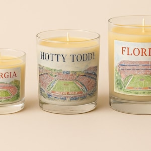 May include: Three glass candles with yellow wax and white labels. The labels feature stadium illustrations and the words "GEORGIA", "HOTTY TODDY", and "FLORIDA" in red and blue. The candles are of varying sizes.
