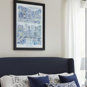 Op de afbeelding: Een slaapkamerinterieur met een marineblauw hoofdbord, wit beddengoed en decoratieve kussens. Een ingelijste prent met blauw-witte architectonische illustraties hangt boven het bed. Een beige sprei voegt textuur toe.