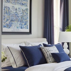 Op de afbeelding: Een slaapkamerinterieur met een bed met witte en marineblauwe beddengoed, aangevuld met decoratieve kussens. Een ingelijste blauw-witte prent siert de muur. Marineblauwe gordijnen omlijsten een raam en een witte lamp staat op een nachtkastje.