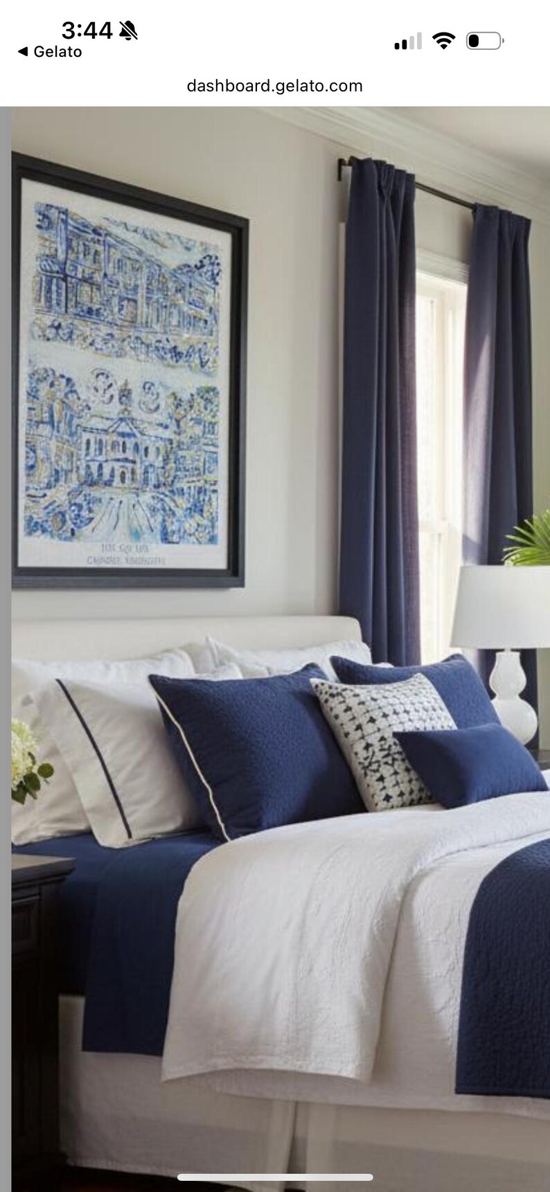 Op de afbeelding: Een slaapkamerinterieur met een bed met witte en marineblauwe beddengoed, aangevuld met decoratieve kussens. Een ingelijste blauw-witte prent siert de muur. Marineblauwe gordijnen omlijsten een raam en een witte lamp staat op een nachtkastje.