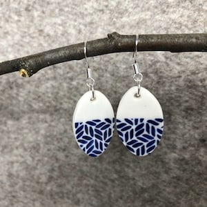 Puede incluir: Un par de pendientes de cerámica blanca con un patrón geométrico azul y blanco. Los pendientes son de forma ovalada y tienen ganchos plateados.