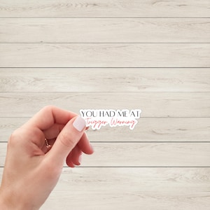 May include: White sticker with the text "You had me at trigger warning" in black font. The word "trigger" is in pink.