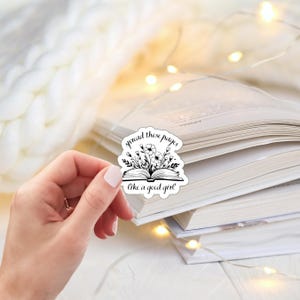 May include: A black and white sticker with the text "spread those pages like a good girl" and a floral book design. The sticker is held in a hand, with a stack of books and fairy lights in the background.
