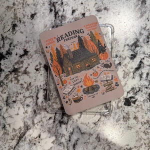 May include: A clear phone case with a decorative insert featuring a cabin scene with the words "Reading Retreat." The insert has a cozy cabin illustration with fall foliage, a campfire, and the text "For the Cozy Readers."