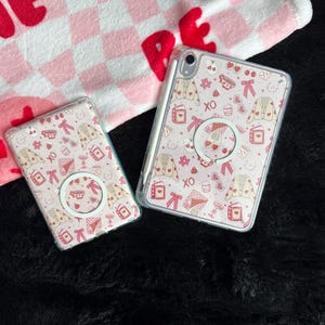 May include: Two phone cases and a tablet case with a pink and white pattern featuring hearts, bows, and clothing. Each case has a clear border and a circular grip. The cases are on a black surface with a pink and white checkered towel in the background.
