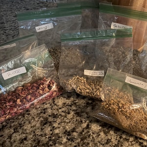 Könnte beinhalten: Eine Sammlung von durchsichtigen, wiederverschließbaren Plastikbeuteln, gefüllt mit verschiedenen getrockneten Kräutern und Blüten. Die Etiketten auf den Beuteln lauten Rose, Echinacea Purpurea, Chamomile, Hibiscus und Raspberry. Die Beutel sind auf einer gesprenkelten Oberfläche angeordnet.