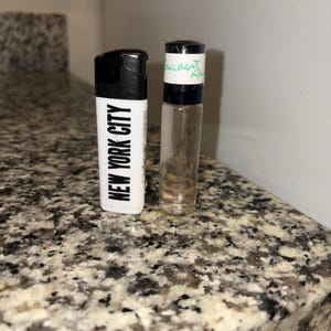 May include: A white rectangular lighter with black text reading NEW YORK CITY and a black top, positioned next to a small clear glass cylindrical vial with a black cap and a white label featuring handwritten green text.