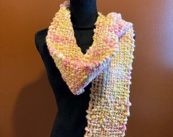 May include: A handwoven scarf in shades of yellow, pink, and white, displayed on a black mannequin. The scarf features a textured weave with fringe at the ends. The color palette is soft and warm, suitable for fall or spring.