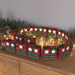 May include: A Christmas train decoration made of red and green boxes, arranged in a circle and lit with fairy lights. The boxes have white accents and the words "Merry Christmas".