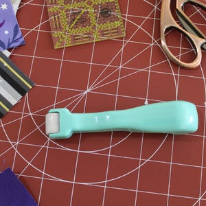 May include: A teal seam roller with a white roller head, positioned on a red cutting mat. The image also shows fabric scraps with Halloween-themed patterns, a clear ruler, and gold scissors.