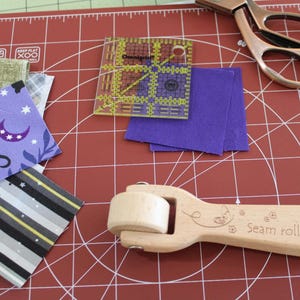 May include: A red self-healing cutting mat with a seam roller, a ruler, and fabric scraps. The seam roller is made of wood and has the words "Seam roller" engraved on it. The ruler is clear plastic with yellow markings. The fabric scraps are purple, black, white, and gray.