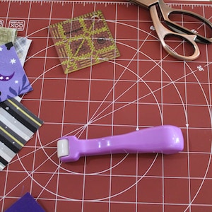 May include: A purple fabric roller tool on a red cutting mat with a ruler and fabric scraps. The ruler has the text "Keep Flat" on it.