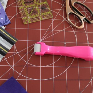 May include: A pink roller tool with a white wheel, used for crafting and sewing projects. The roller is on a red cutting mat with a grid pattern.