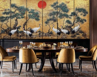 Op de afbeelding: Een ronde houten eettafel met zes stoelen. De stoelen zijn bekleed met een gouden stof en hebben zwarte poten. De tafel is gedekt met couverts, waaronder glazen, bestek en servetten. De muur achter de tafel heeft een groot, verguld scherm met een schilderij van kranen en dennenbomen.