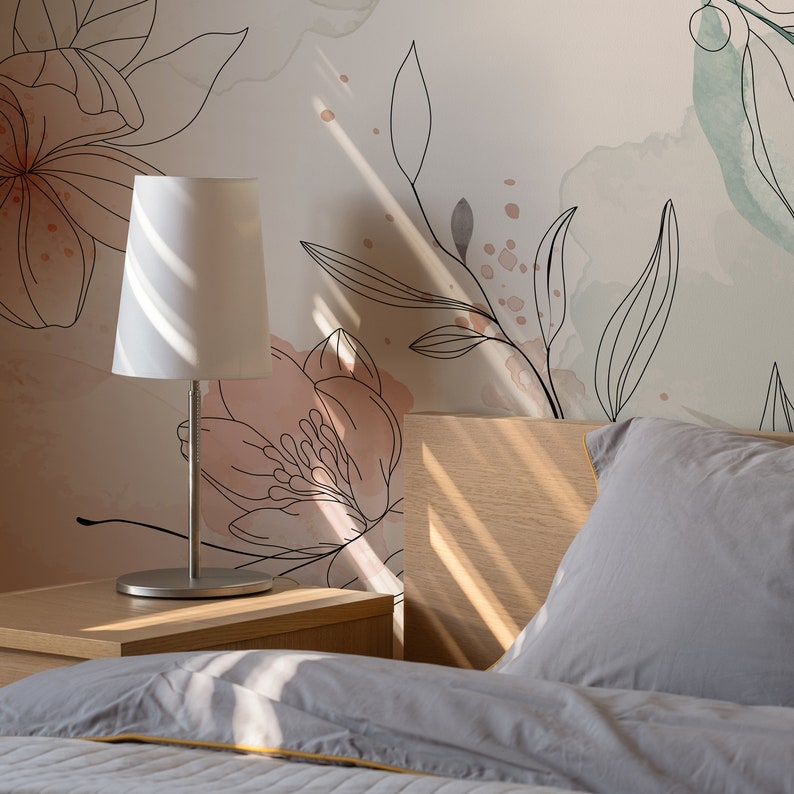 Pu&ograve; includere: Una lampada da comodino bianca con un paralume bianco si trova su un comodino in legno accanto a un letto con biancheria da letto grigia. La parete dietro il letto presenta un design di carta da parati floreale con accenti di acquerello rosa e verde.