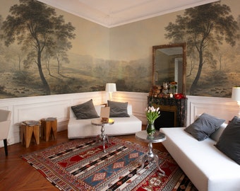 Könnte beinhalten: Ein Wohnzimmer mit weißen Sofas und grauen Kissen. Der Raum hat eine weiße Wandverkleidung und ein Wandbild mit einer Waldszene an den Wänden. Auf dem Boden liegt ein Teppich mit einem rot-blau-weißen Muster.