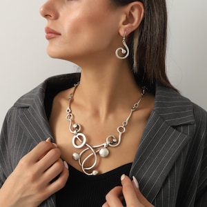 Peut inclure: Un ensemble de collier et boucles d'oreilles argentés. Le collier présente un design abstrait avec des cercles, des sphères et des lignes courbes. Les boucles d'oreilles ont un design en spirale. Les bijoux sont portés avec un haut noir et un blazer à rayures grises.