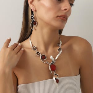 Könnte beinhalten: Silberfarbenes Halsketten- und Ohrring-Set mit roten Edelstein-Akzenten. Die Halskette hat ein einzigartiges, fließendes Design mit Spiral- und blattartigen Elementen. Die Ohrringe haben ein ähnliches Design mit einem einzelnen roten Stein.